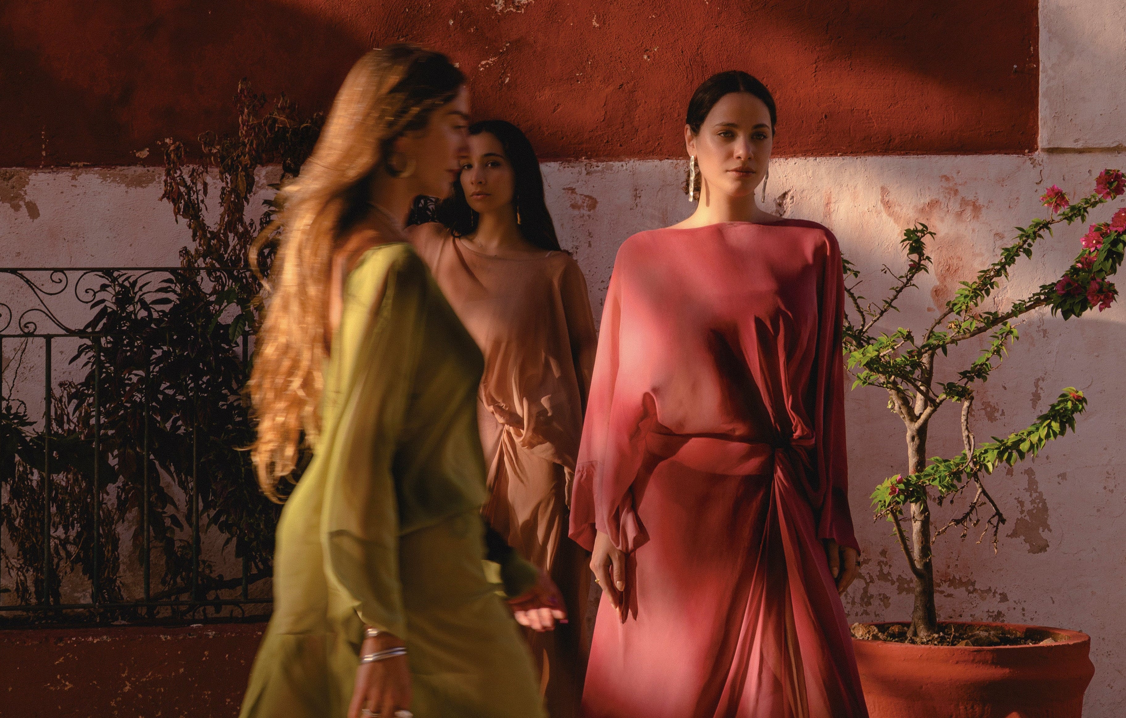 Three women in flowing resort dresses walking on a sunlit street with a rustic red wall